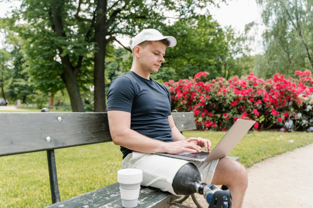 An adult male with a prosthetic leg working on a laptop while sitting on a bench in a park.